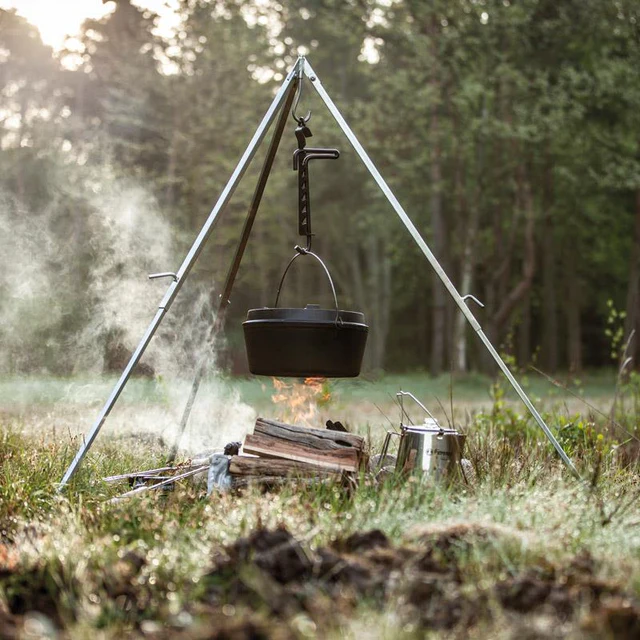 ein brennendes Lagerfeuer unter einem Dreibein mit einem daran hängenden großen Dutch Oven auf einer Wiese vor einem Wald