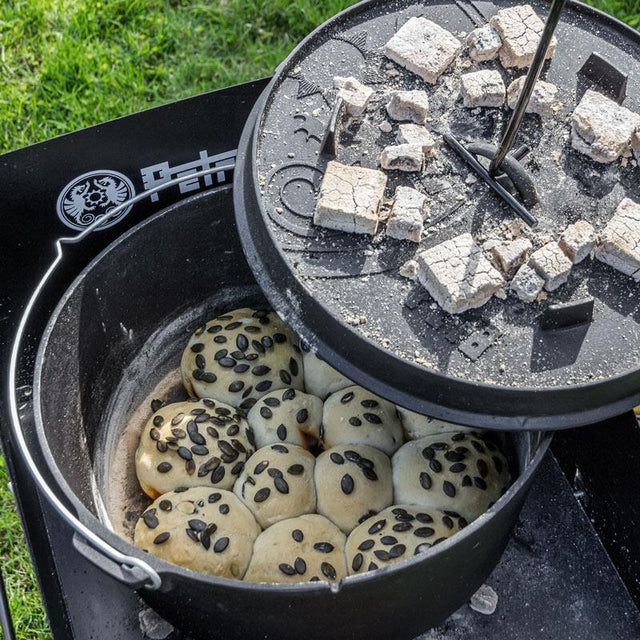 gebackene Kürbiskernbrötchen in einem Dutch Oven auf einem Feuertisch, bei dem mit einem Deckelheber der Deckel weggehoben wird