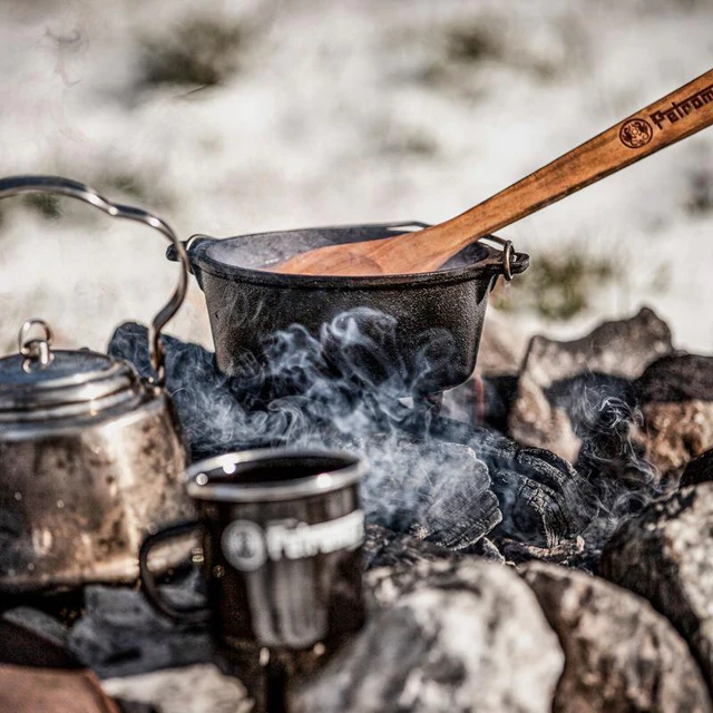 ein Gusstopf mit Holzlöffel, eine Emailletasse und eine Teekanne auf rauchender Kohle im Schnee