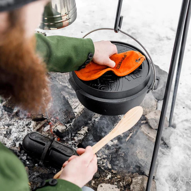 mit einem topflappen packt ein bärtiger Mann an den Deckel eines gusseisernen Dutch Oven, der an einem Haken über einer Feuerstelle hängt